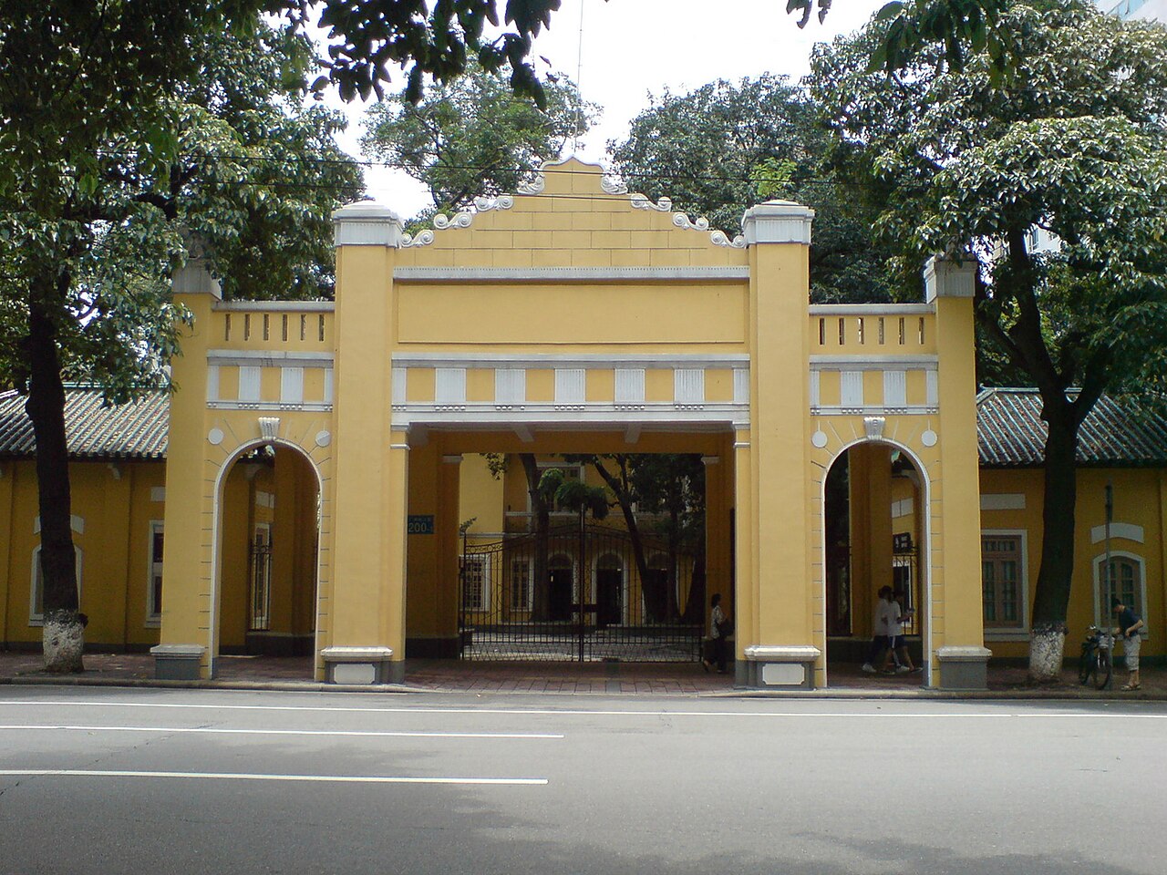 A yellow building with a decorative archway entrance and white accents, surrounded by trees, appears to be related to th