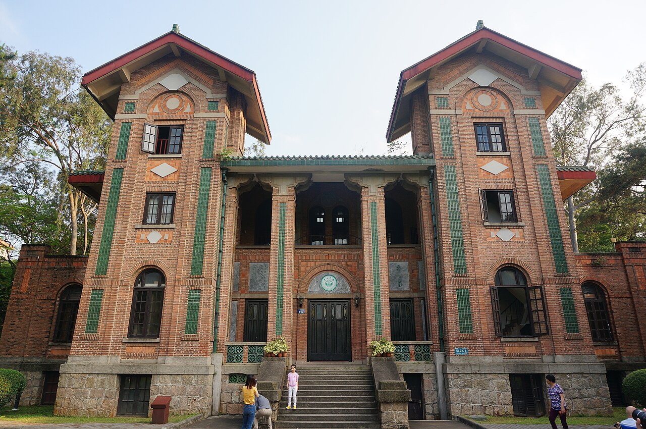 A brick church campus with two towers, featuring green decorative tiles, and people standing on the steps and nearby, su