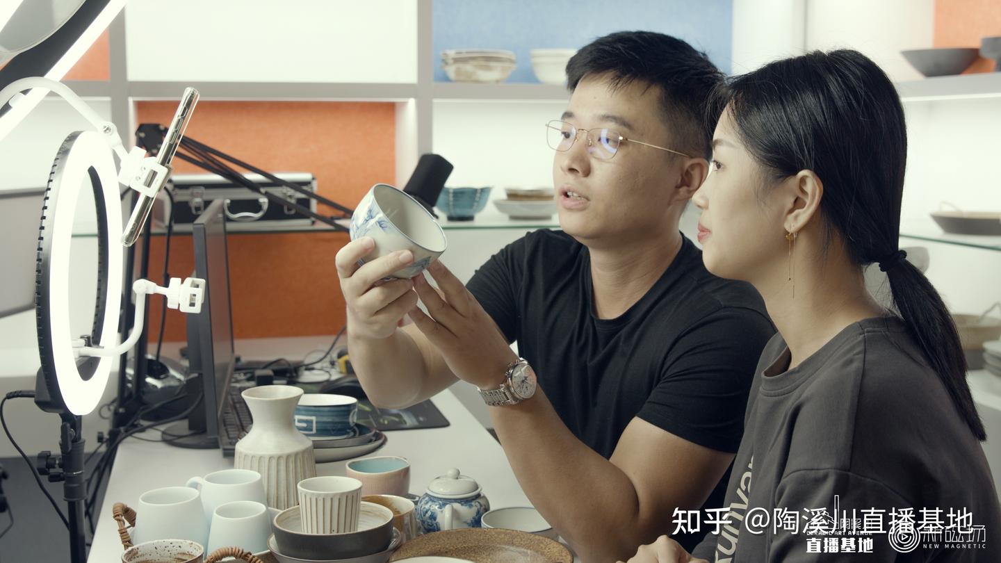 景德镇陶瓷直播间的现场场景，主播在堆满瓷器的背景前向手机镜头介绍产品