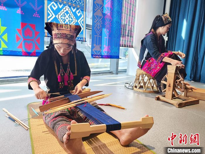 传习所教学区场景，学员在老织工指导下练习腰织机操作，墙上可见传承人简介展板，男性学员也在其中