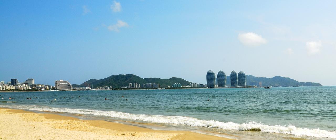 三亚湾的海滩与城市天际线：椰树列植的海岸线上，人群散布在金色的沙滩上，后方是天际线轮廓和三面环山的城市格局