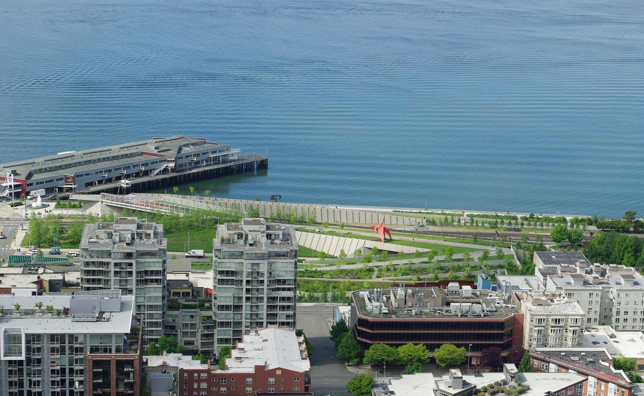 从 Space Needle 高位俯拍的 Olympic Sculpture Park：Z 形平台从城市端的 PACCAR Pavilion 跌落到 Elliott Bay 水边，跨过 Elliott Avenue 与 BNSF 铁路两道线性基础设施
