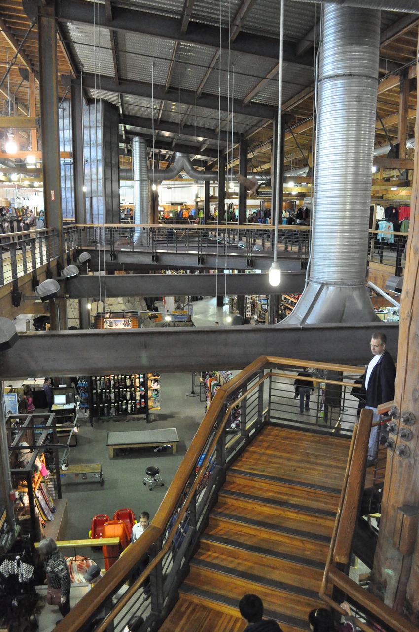 REI 旗舰店内部 galleria 与多层零售空间，画面背景可见攀岩柱玻璃罩内一侧