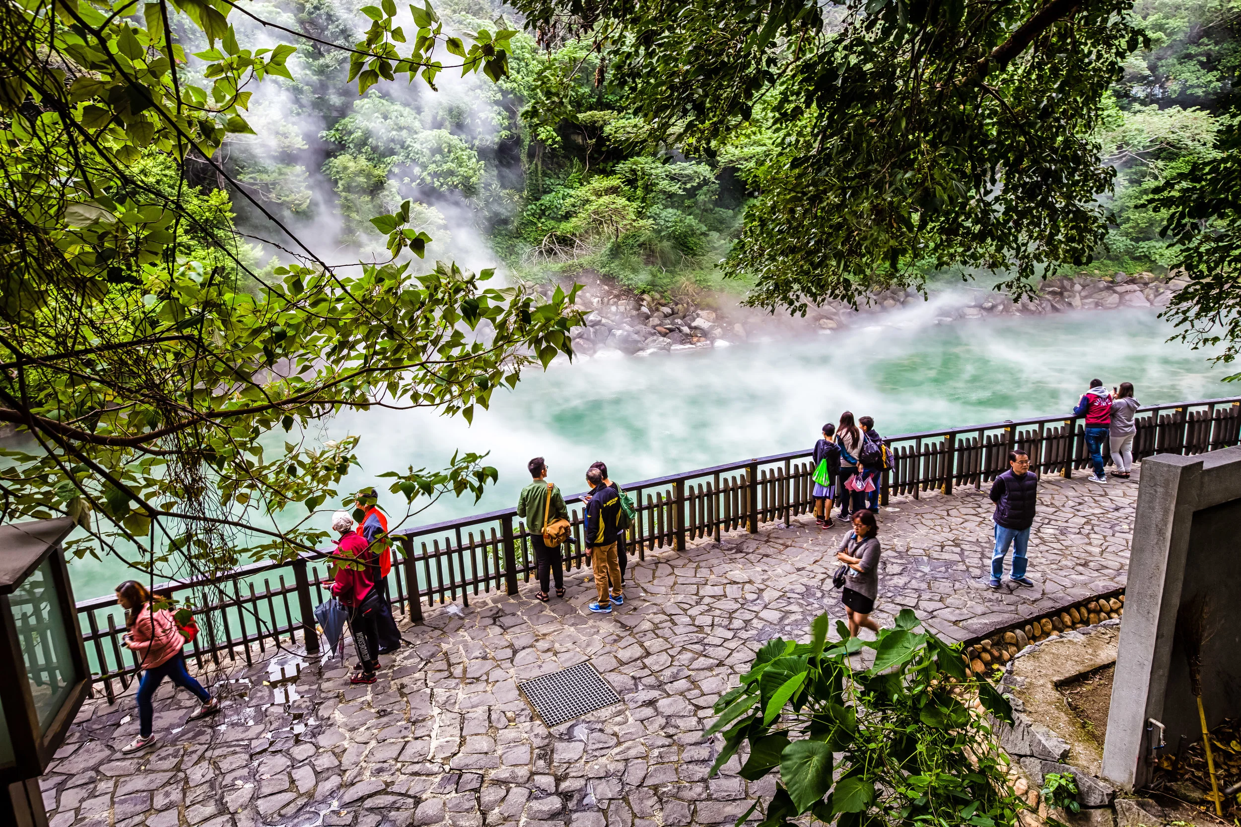 People are gathered along a railing overlooking steaming, turquoise geothermal w