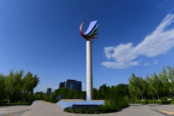A tall pole with a colorful abstract sculpture on top stands in front of a park 